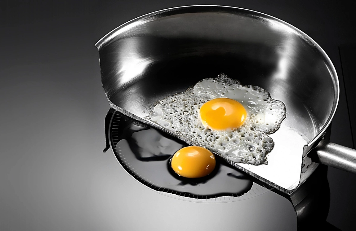 cutaway view of an egg frying in a pan on an induction hob. One egg is cooking in the pan, another is sitting uncooked on the hob ring
